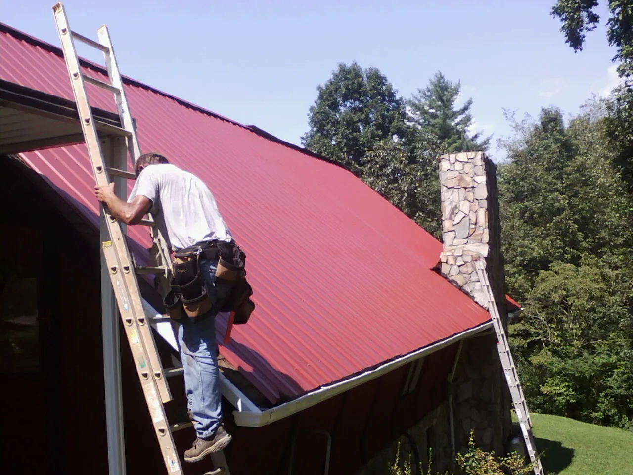 Roofer on a ladder beside a red metal roof and stone chimney — Soffit & Fascia Repair by Roofing Brothers in Mountainside
